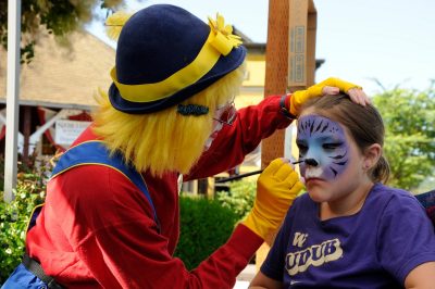 A clown with yellow hair, blue hat, and a red shirt paints a purple & blue tiger onto the face of a young lady.