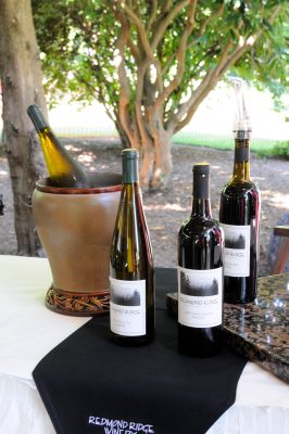 A table with white and black tablecloths display bottles of wine under the shade of trees. The wine is labeled Redmond Ridge, and the black tablecloth says Redmond Ridge Winery.