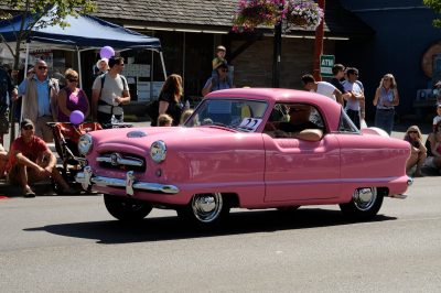 A pink automobile travels down Railroad Ave during the Snoqualmie Railroad Days parade. People watch from the street edge.