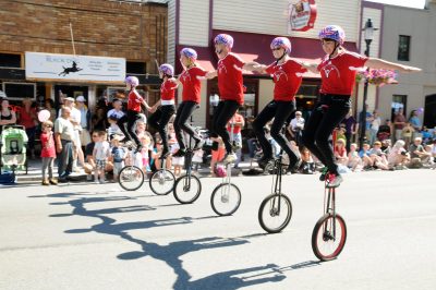 6 people riding tall unicycles with outstretched arms linked ride down Railroad Ave during Snoqualmie Railroad Day's parade. Visitors line the street edge watching the parade.