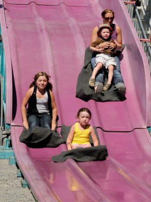 Two kids and a parent/child pair sitting on fabric sheets slide down a purple carnival slide