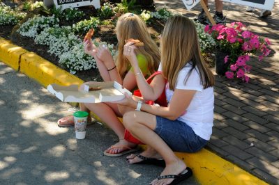 Two people sitting on a curb watch the Parade. Both hold pizza crusts, with one holding an empty pizza box.