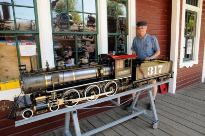 Russ N. displays his large live steam locomotive next to the Snoqualmie depot. The 4-6-0 locomotive rests on a rolling gray frame.