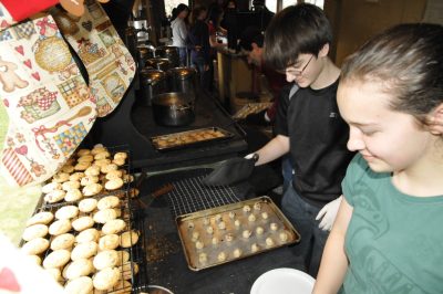 Two young volunteers stand in front of an old Army range stove. Cookies on racks stand on the back, and a tray with cookie dough sits on the stove top. In the background, another volunteer pulls a cooked tray of cookies out of another stove.