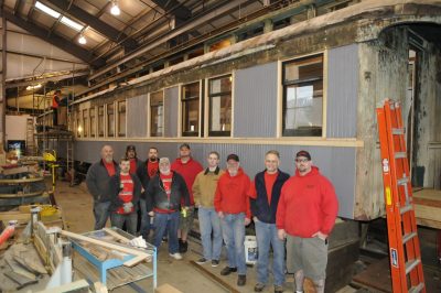 Members of The Ancient and Honorable Order of E Clampus Vitus pose in front of Chapel Car Messenger of Peace in the CRW. The car has siding on it in primer gray. More volunteers stand on scafolding further back, working on the roof.