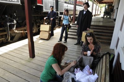 Two actors stand ready for action along the boardwalk of the Snoqualmie depot. One is in a porter's outfit and has a cart with luggage aboard near the railroad tracks. The other is in an old-fashioned suit holding a suitcase near the baggage room doors. Between the actors is a movie crew member with radios and listening to a headset. Two other crew members attend to costumes on a bench. US Plywood 11 sits at the platform with the Silver Bullet and 4024 behind it.