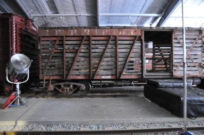 Northern Pacific Stock Car #83296 sits in the Train Shed prior to filming for "You Can't Win". A spotlight is set up next to the car, and a pad is set on the ground at the doorway.