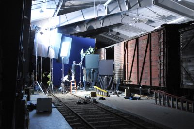 One of the former GN automobile boxcars rests in the Train Shed. A blue screen is set up outside the open doors, with lights and reflectors set up around. Film crew are present, helping filming.