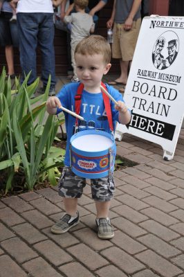 A young visitor plays with a Thomas drum on the brick walkway at the Snoqualmie Depot. Other visitors can be seen in line behind him.