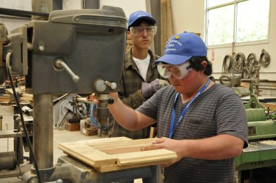 A 2012 RailCamper drills a hole using a drill press in the CRW. Another RailCamper looks on.