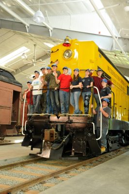 Twelve teens stand on the front platform of Weyerhaeuser #1 in the train shed. All are wearing blue baseball hats as part of RailCamp 2012.