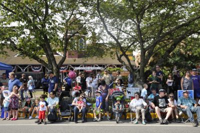 Many people line Railroad Ave. in Snoqualmie for Snoqualmie Railroad Days 2012. Some are sitting on the curb, others in camp chairs, while more stand on the sidewalk. Behind them stand two small trees, through which the Snoqualmie Depot can be seen.