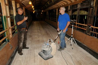 Two workers talk within SP&S 218. One holds a handle of a floor sander.