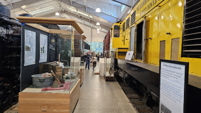 A view down one of the Train Shed aisles, to the left is a black steam locomotive. On the right is a bright yellow diesel locomotive.