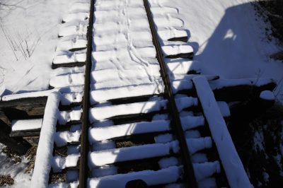 Railroad tracks in the snow. The rails are uncovered, but the ties are covered. The picture shows the change from the grade to a bridge, with coyote tracks in the snow.