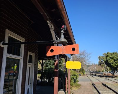 The train order board at Snoqualmie Depot in the "Permissive" aspect as seen from the platform.  The board is perpendicular to the tracks with a yellow board hanging, and the red lights are displayed.