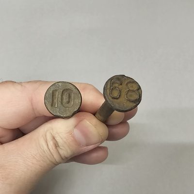 Two railroad date nails used to mark what year they had been produced. Both are held in a hand. 10 is imprented into the top of the left nail, and 68 is raised on the right nail.