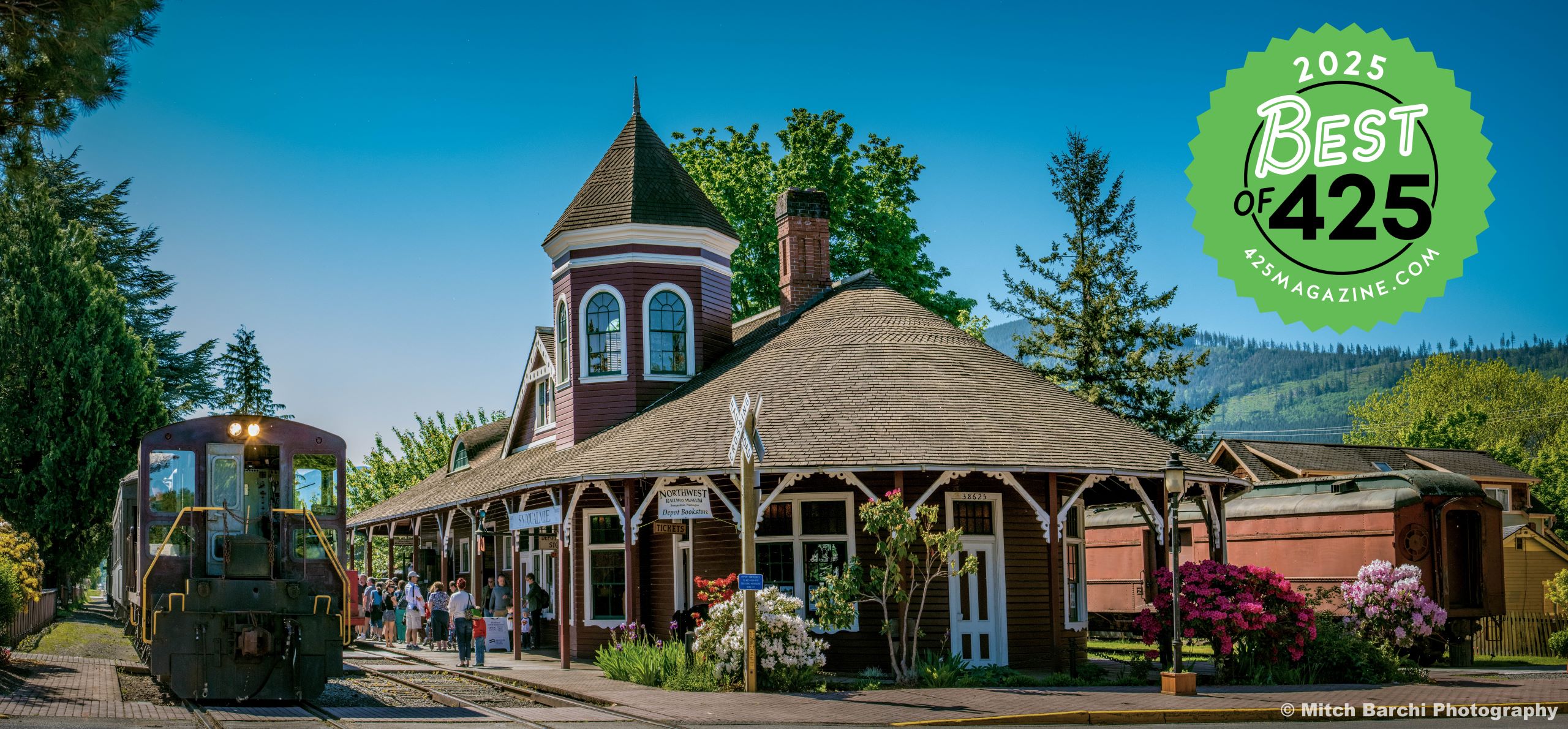 A large red building with a turret sits in the sun. Passengers mill in front, and a burgundy locomotive with a train waits on the tracks. Other train cars can be seen behind the building.