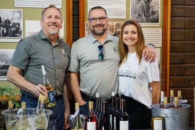 Three people stand behind a table full of bottles of wine.