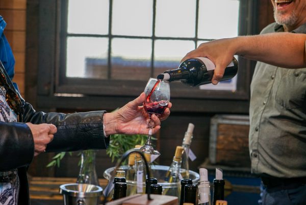 A person to the left holds a wine glass, which is filled from the right. Below are several wine bottles on a table. In the background, an old window lets in light.