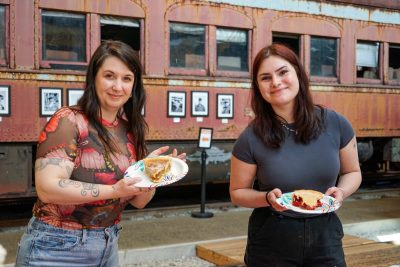 Two ladies stand before an old railcar displaying slices of pie on plates. One has an amber filling, the other is red.