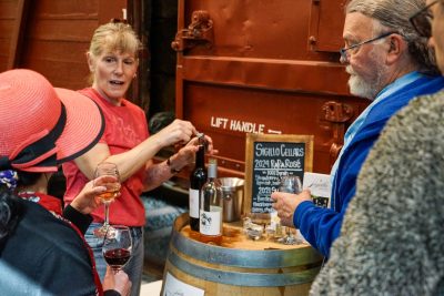 A server from Sigillo Winery chats while opening a new bottle of wine. the bottle and a sign are resting atop a wine barrel.