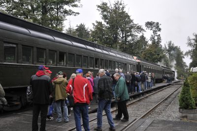 People standing in line next to old green steel passenger coaches. Steam from a steam locomotive can be seen from the front of the train.