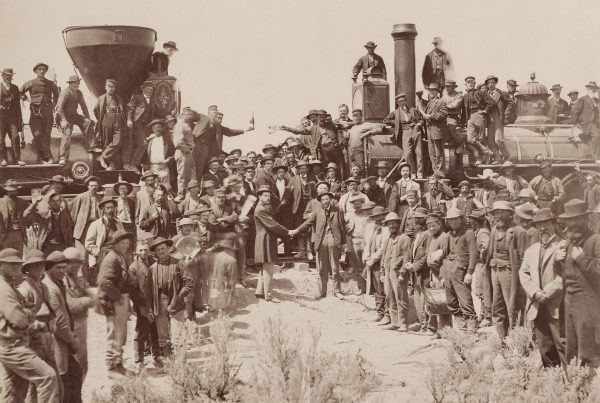 A crowd of people celebrating for a photographer with two steam engines facing nose to nose. Two men on the ground are shaking hands and two leaning off the front of the locomotives are holding champagne bottles. Taken 1869.
