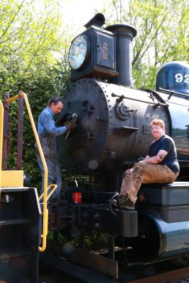 Two men are standing in front of a steam locomotive. One has a wrench and is standing in front of the smokebox door, wrench in hand. The other is sitting on the cylinder cover, smiling at the camera.