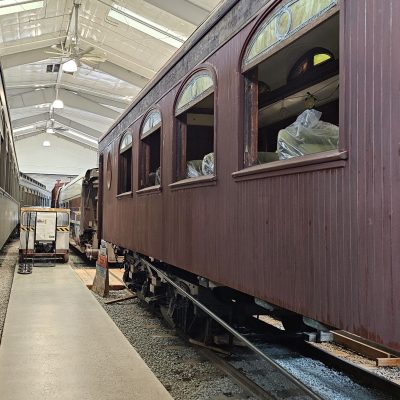 A brown wooden train car inside a building. Green seats wraped in plastic can be seen through the open window frames.