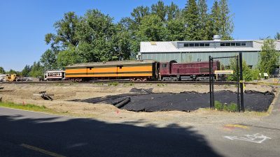 New construction at the RHC. Trees have been cut down and a hole dug for a parking lot expansion. A purple locomotive with a green and orange baggage car are seen sitting on tracks, with the Train Shed behind them.