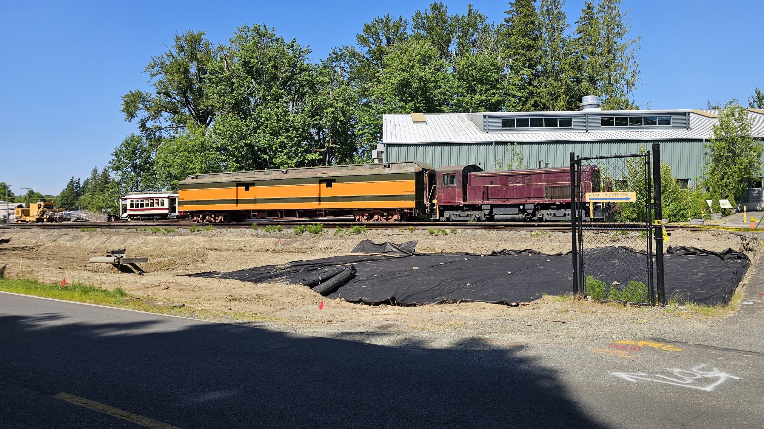 New construction at the RHC. Trees have been cut down and a hole dug for a parking lot expansion. A purple locomotive with a green and orange baggage car are seen sitting on tracks, with the Train Shed behind them.