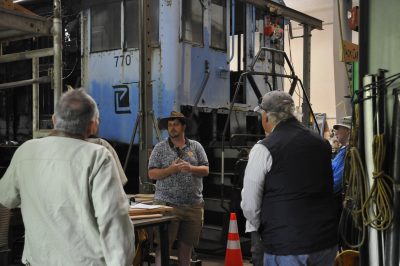 A person stands in front of a blue and black diesel locomotive inside a workshop. The person is talking to several other people around them on a tour