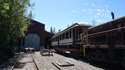 A purple locomotive pushes a wooden train car body on flatcar. Ahead is a purple building. the door in front of the car is open.