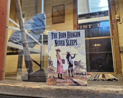 The book "The Iron Dragon Never Sleeps" stands in an open doorway with plexiglass behind it. Visible behind the doorway is the interior of the Bunk Car, a converted Boxcar for living space.