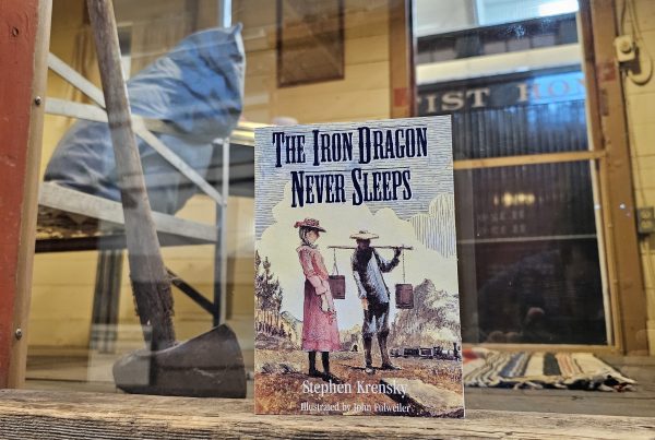 The book "The Iron Dragon Never Sleeps" stands in an open doorway with plexiglass behind it. Visible behind the doorway is the interior of the Bunk Car, a converted Boxcar for living space.