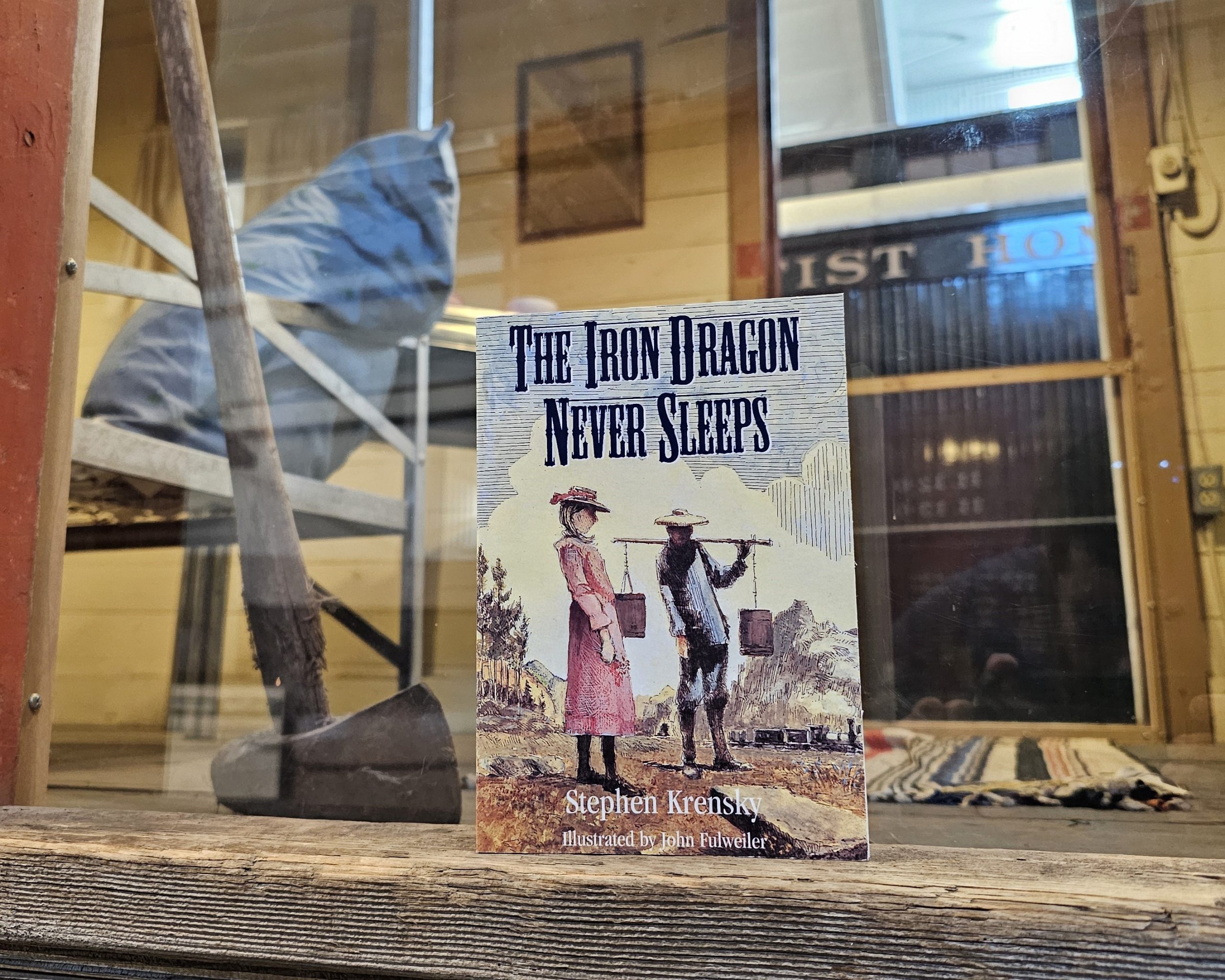 The book "The Iron Dragon Never Sleeps" stands in an open doorway with plexiglass behind it. Visible behind the doorway is the interior of the Bunk Car, a converted Boxcar for living space.