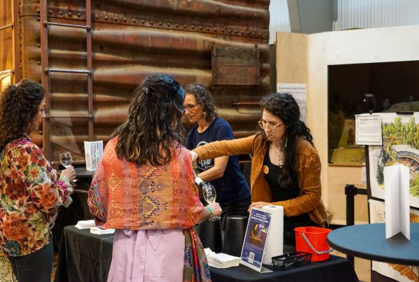 Two women pour wine in front of a model train exhibit and a refrigerator car. Two visitors chat with them.