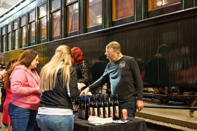 A man pours wine in front of a green passenger car. Two ladies watch over the table loaded with wine bottles at the wine being poured.
