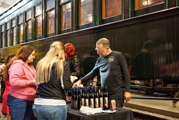 A man pours wine in front of a green passenger car. Two ladies watch over the table loaded with wine bottles at the wine being poured.