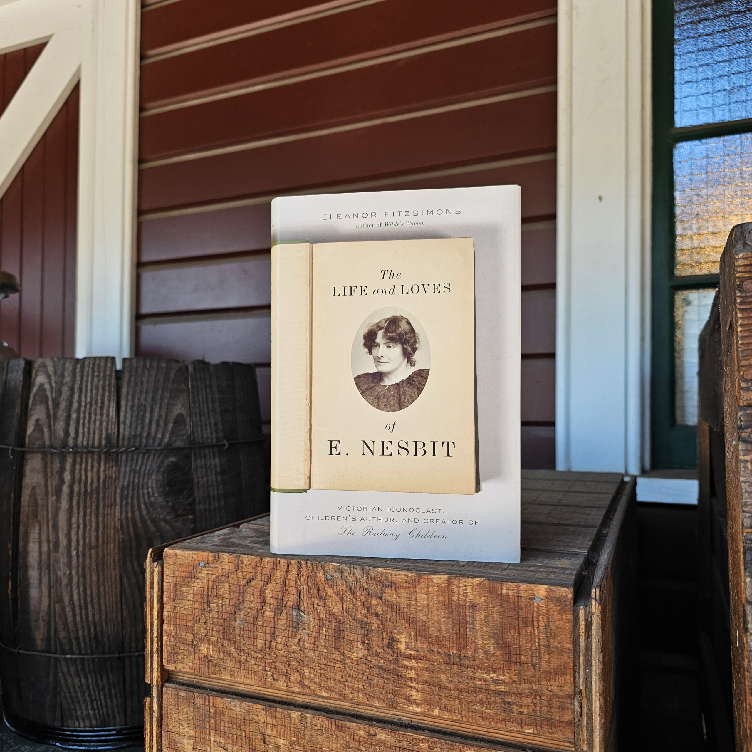 A book title "The life and Loves of E. Nesbit" displayed atop a wooden box. An old barrel is to its left, and the side of the Snoqualmie Depot is behind it.