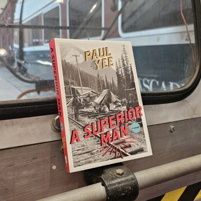 The book "A Superior Man" by Paul Yee rests on the front of a motor car. Through the windows can be seen the Museum's Amtrak Talgo Bistro Car and Northern Pacific Tool Car