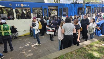 People stand on a sidewalk next to TriMet MAX Type 1 LRV 124. Some sign messages, some take photos, others talk. One person wears a Type 1 costume.