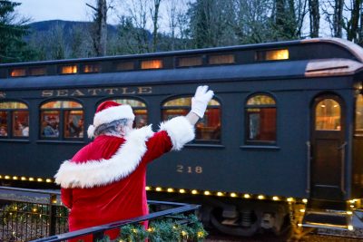 A green wooden passenger car rolls by loaded with Santa Train passengers. Holiday lights illuminate the bottom of the car. Santa waves to the train from a deck close to the tracks.