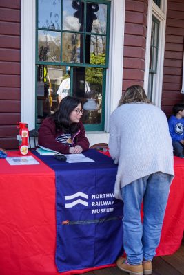 Oral History intern Mayna sits at a table in front of the Snoqualmie Depot's bay window. The table is covered by a red tablecloth with a blue runner featuring the Museum's logo and train. Mayna is helping a visitor fill out a form.
