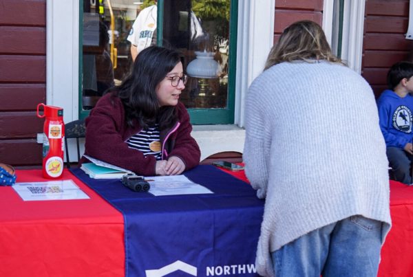 Oral History intern Mayna sits at a table in front of the Snoqualmie Depot's bay window. The table is covered by a red tablecloth with a blue runner featuring the Museum's logo and train. Mayna is helping a visitor fill out a form.