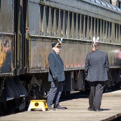 Two train crew members with bunny ears chat on the platform at the RHC. The passenger train sits behind them, and there is a yellow step box next to one of the crew.