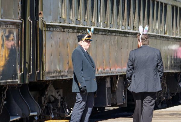 Two train crew members with bunny ears chat on the platform at the RHC. The passenger train sits behind them, and there is a yellow step box next to one of the crew.