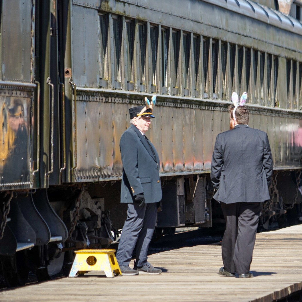 Two train crew members with bunny ears chat on the platform at the RHC. The passenger train sits behind them, and there is a yellow step box next to one of the crew.