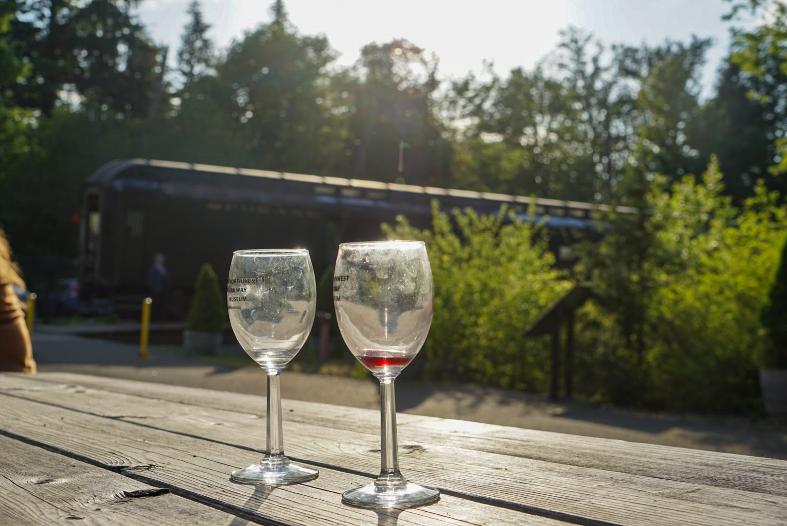 Two wine glasses glint in the sun atop a picnic table. The Museum's logo can be seen on the glasses, and a small amount of wine is left in one. The Museum's train can be seen in the background waiting.